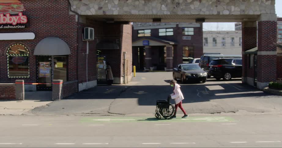 An elderly woman pushes an empty wheelchair along a street