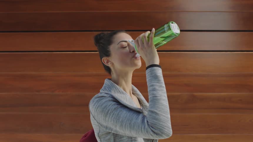 A woman drinking from a green water bottle while standing near a wooden wall
