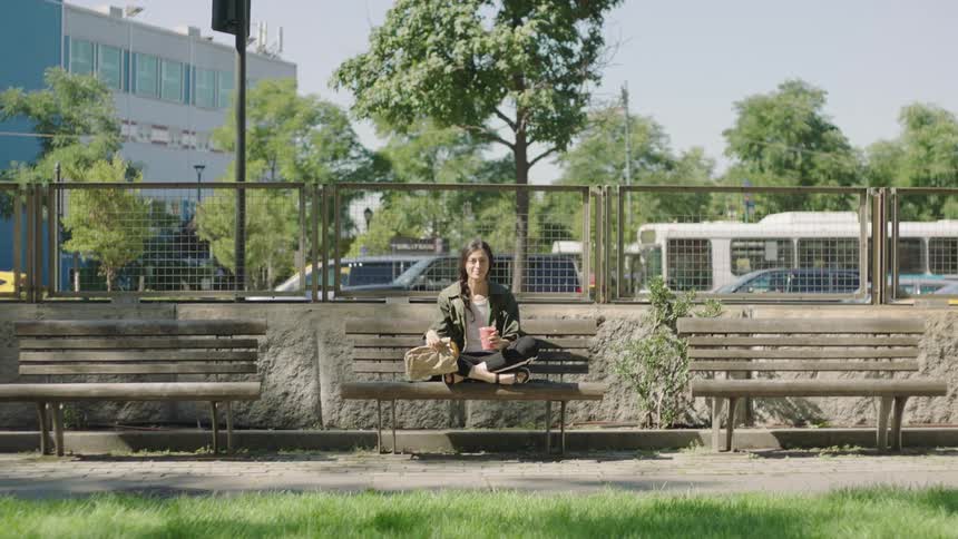 A person sits on a bench with a drink and a purse