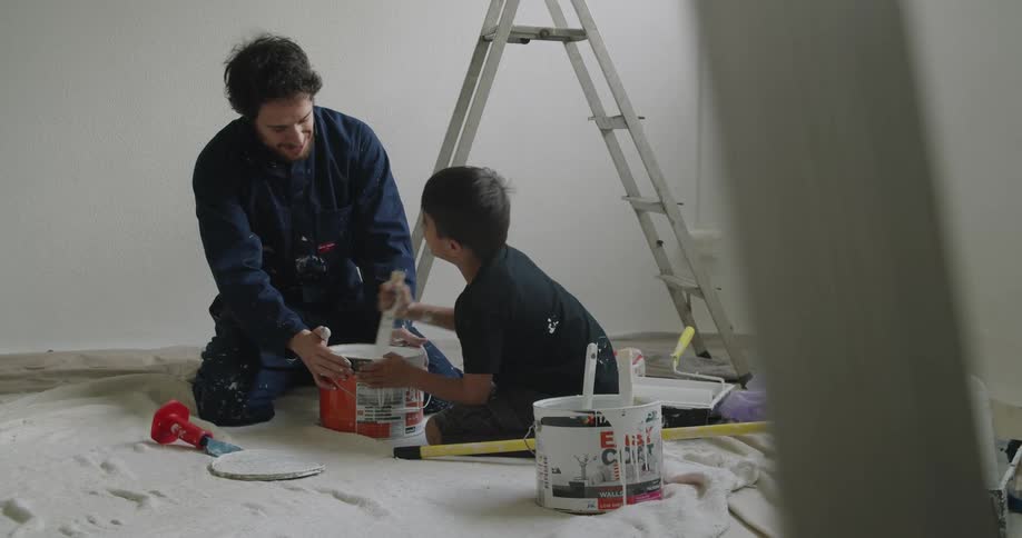 A dad holds the paint can as his young son stirs it with a stick
