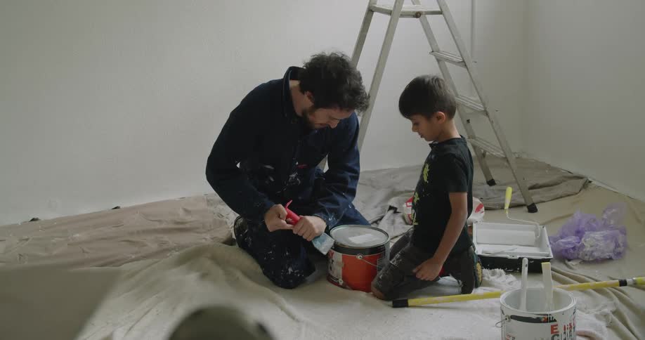 A man shows his son how to open a bucket of paint as they prepare to renovate a room