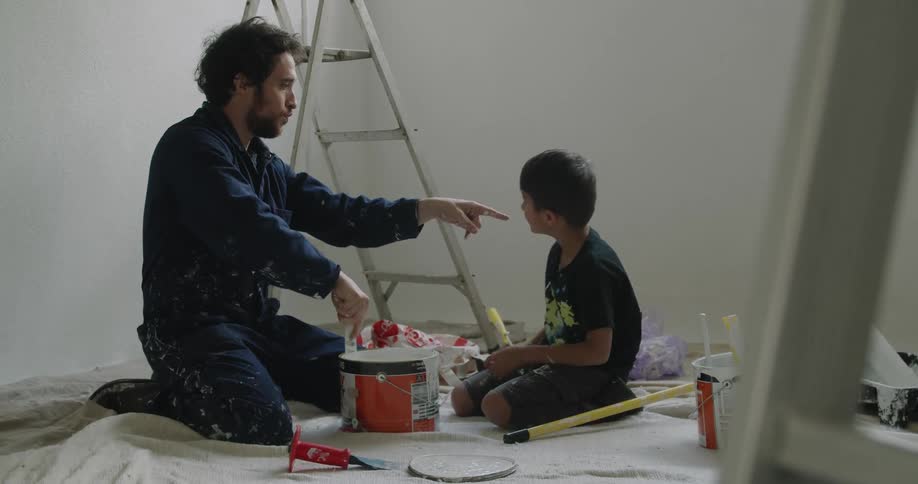 A man mixes a bucket of paint in a room as his son helps out