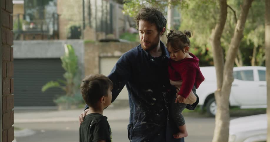 A man holds an infant daughter and fixes the hair of his young son while outside with their dog
