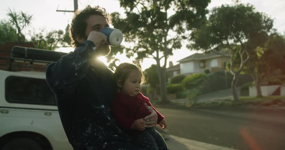 A man holds a female toddler outside in his driveway by the road drinking from a coffee mug