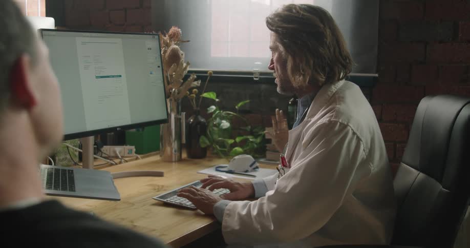 A man in a lab coat takes notes on a computer while talking with another man