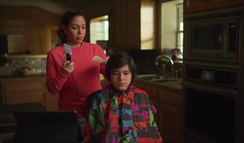 A mom looks at a tablet for instruction while holding clippers and a comb in her hand then she cringes as she starts to cut her nervous sons hair