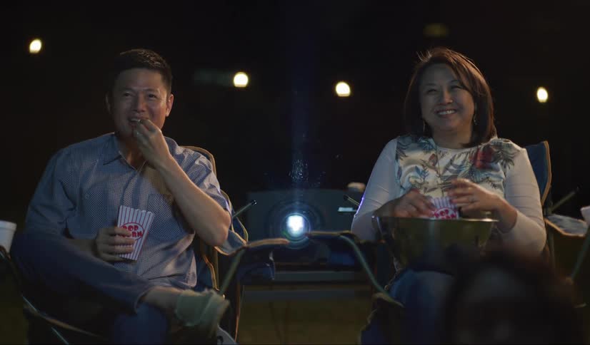 A man and woman watch a movie in the backyard with a projector, eathing popcorn