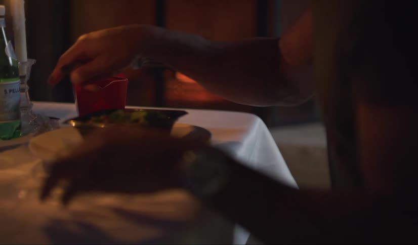 A black man sitting at an outdoor table lit by candle light and a glowing fire in a fireplace gestures at the food in front of him