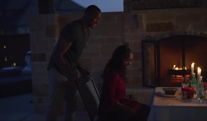 A black man hugs his wife in appreciation after she presents the candlelit table she has set up by the outdoor fireplace