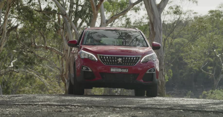 A woman drives a red crossover vehicle down a narrow road near a forest