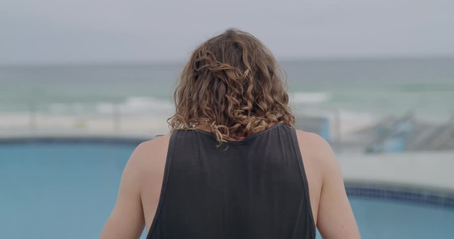 A young man with curly brown hair stands in front of an empty swimming pool