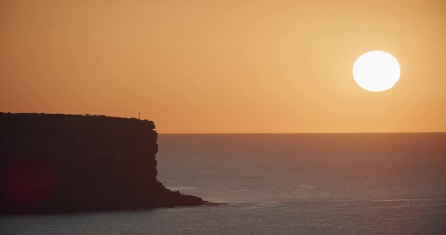 Large sun glows over ocean against orange sky turning cliff into a silhouette