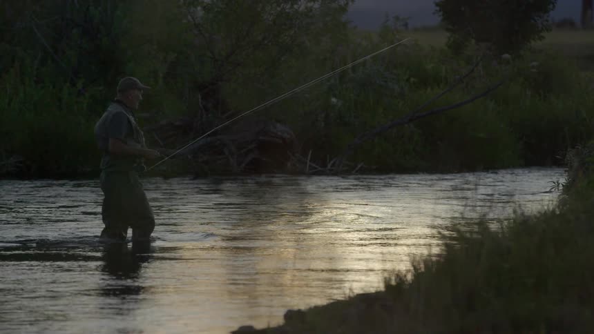 A man with a fishing pole walking across a river at dusk or dawn