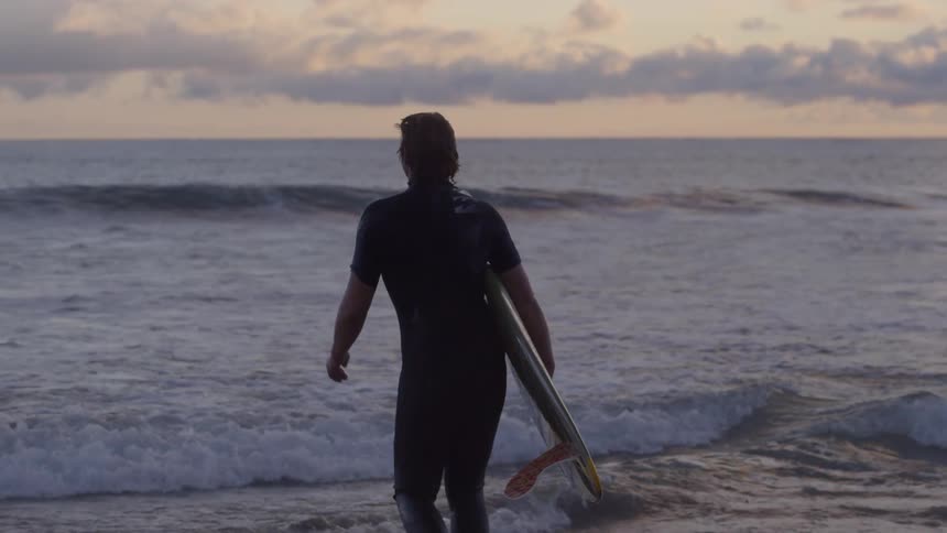 A man carrying a surfboard strides into the ocean at dusk or dawn