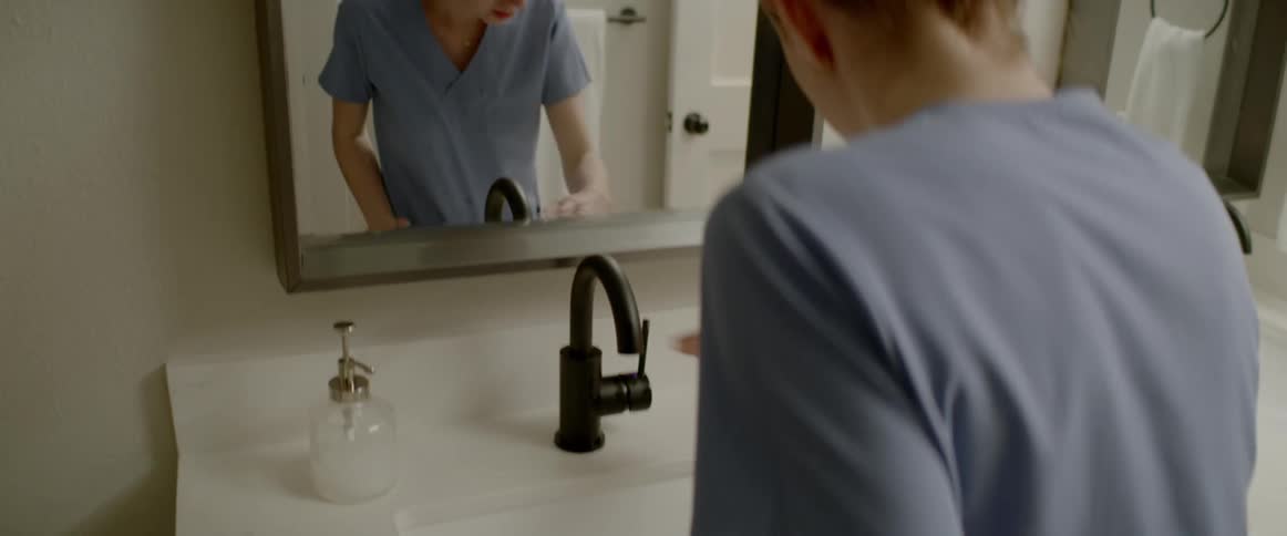 A woman in a scrub top turns on the bathroom faucet and washes her face then towel dries and takes a deep breath