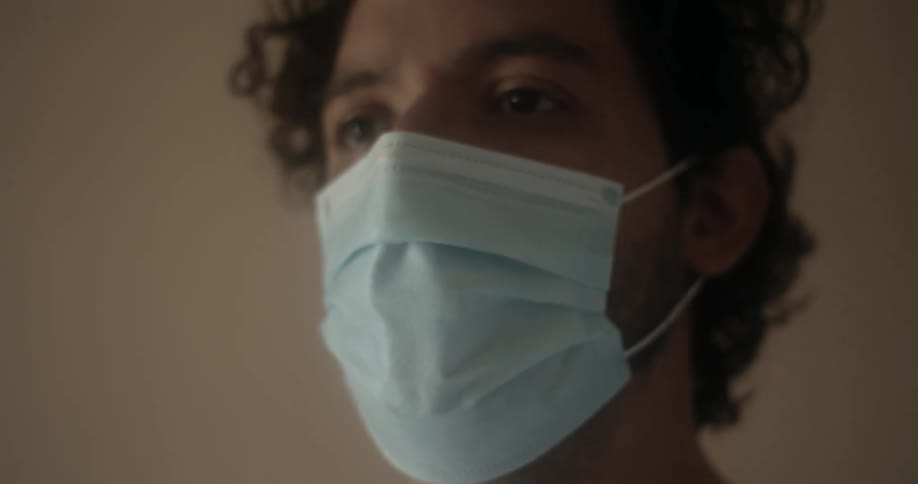A man with curly black hair wears a blue disposable face mask