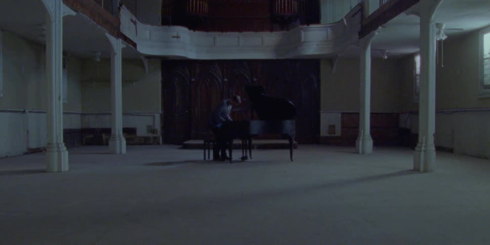 A person stands in a dark room and then sits at a piano