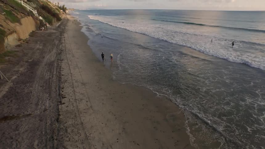 Drone footage of people carrying surfboards and walking along the shoreline as waves roll onshore