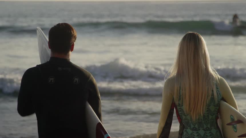 Couple carry surfboards while watching a surfer catch a wave
