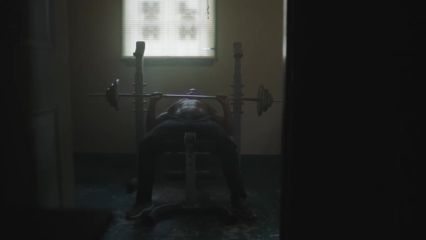 A shirtless man lifts free weights on a bench in a small room with a window