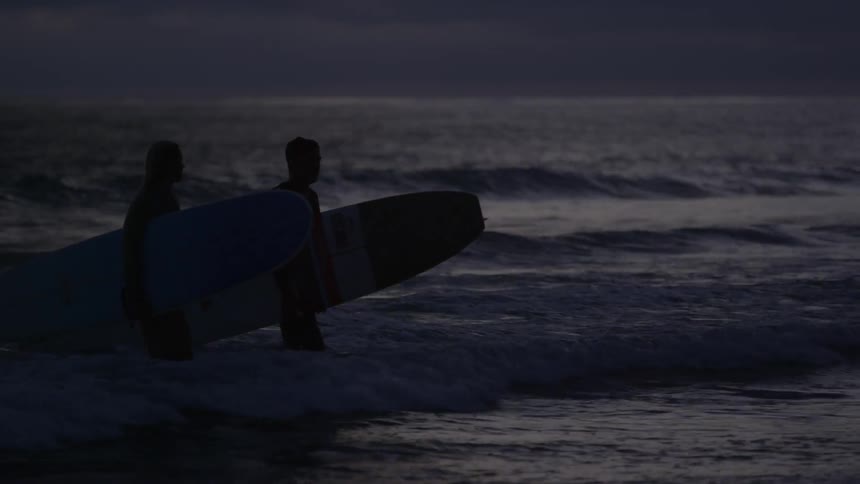 A man and woman walk out of the ocean carrying surfboards