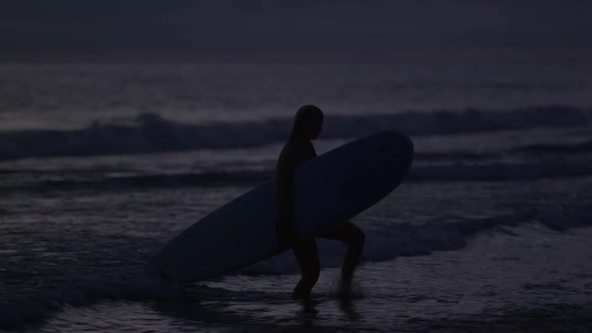A couple walks with surfboards out of the water as night approaches
