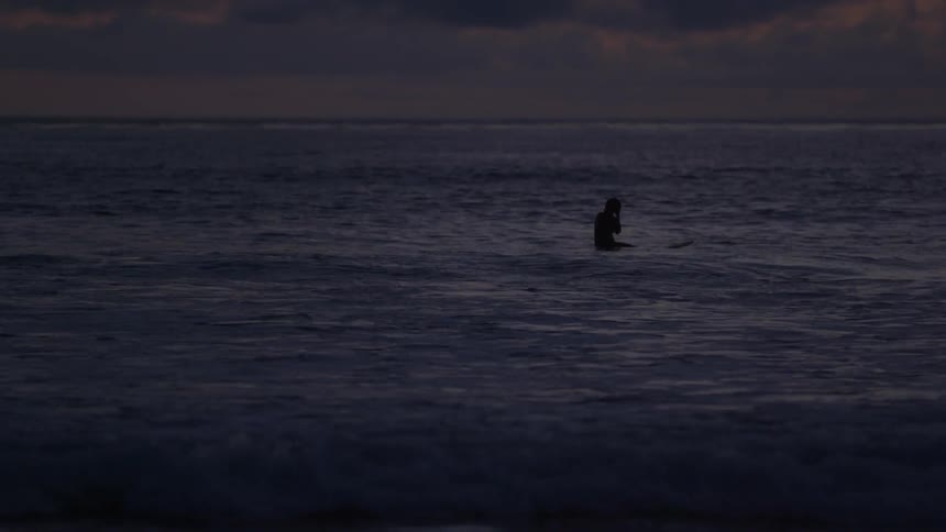 Person sitting on surfboard riding gentle waves as sun sets