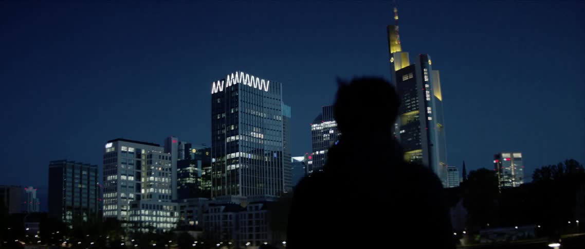 A mans silhouette stands looking at the distant tall buildings and a skyscraper at night
