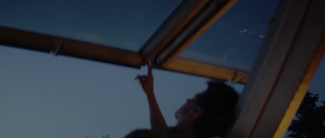 A man leans against the edge of an open skylight reaching up to touch the raised glass while looking out at the sky
