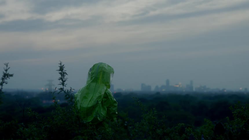 A person in a hazmat suit stands and looks at the city skyline in the distance