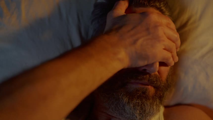 A man lying in bed rubs his face and eyes trying to wake himself up while holding his phone up