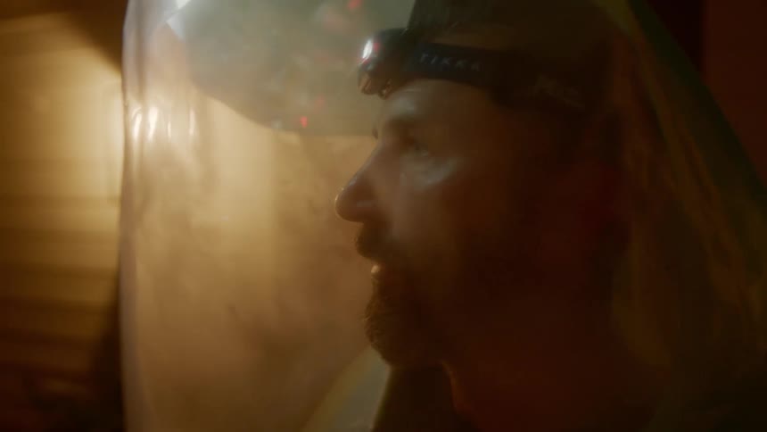 A man wearing a hazmat suit and a headlamp walks out of his house onto the front porch and looks around