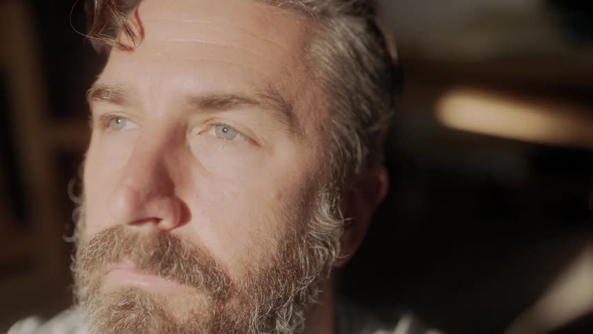 Bearded man looking out the window smiles while he thinks about something