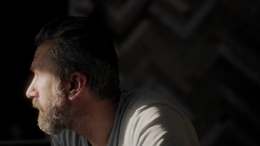 A man sitting before a window indoors and looking through it as sunlight shines on his face