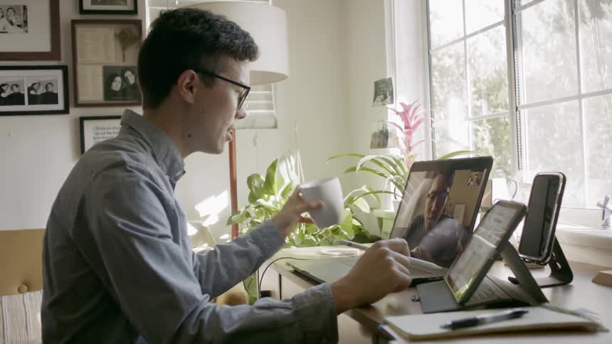 Man drinks and works on tablet while communicating with another over video call