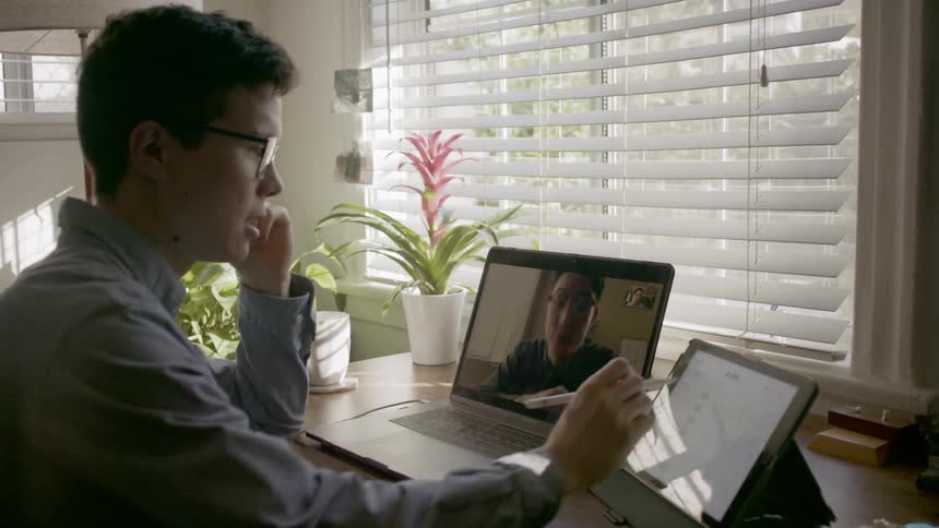 A person talking to another man on a laptop using video chat while operating a tablet with a stylus