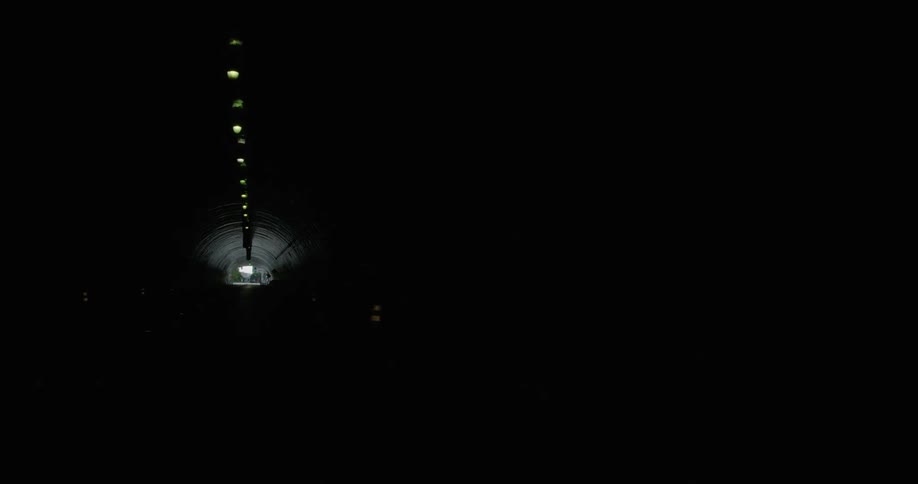 Moving through a train tunnel with lights in a single row on the ceiling towards daylight