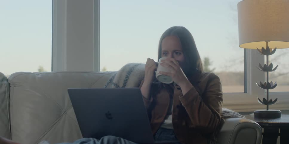 A woman is sitting on the couch drinking from a mug and using her laptop