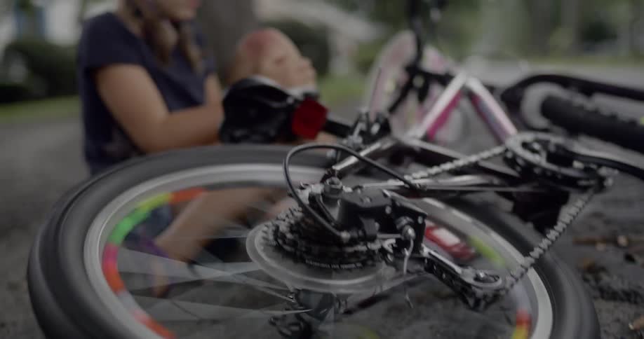 Girl sits on road after fall from bicycle frowning while examining scrape