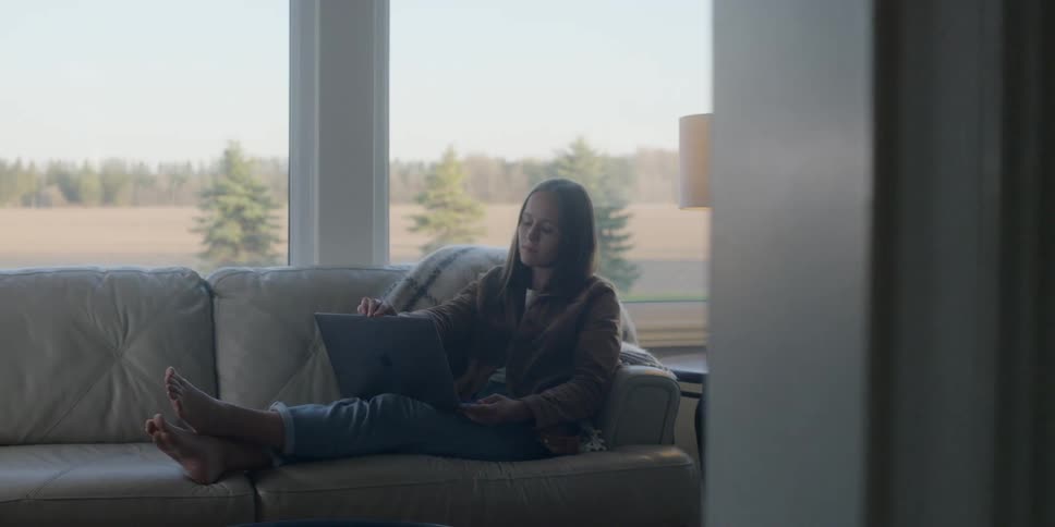 Woman sits back on couch working on laptop