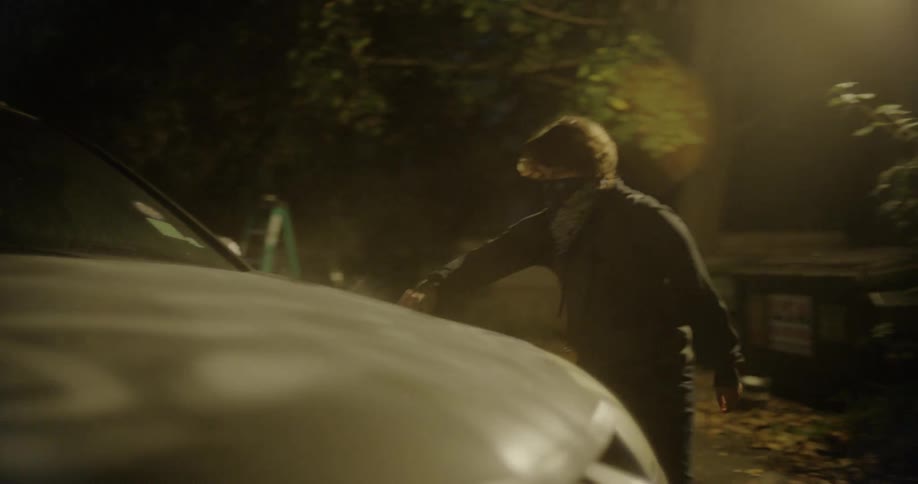 A person wearing a bandana over their face vandalizing a vehicle with spray paint at night