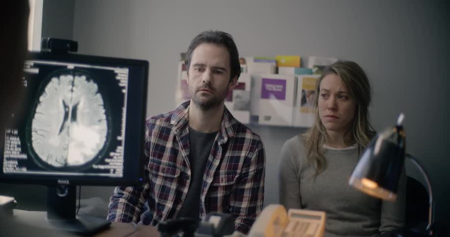 A couple sits in a doctors office as the doctor delivers bad news to them with a brain scan of his display