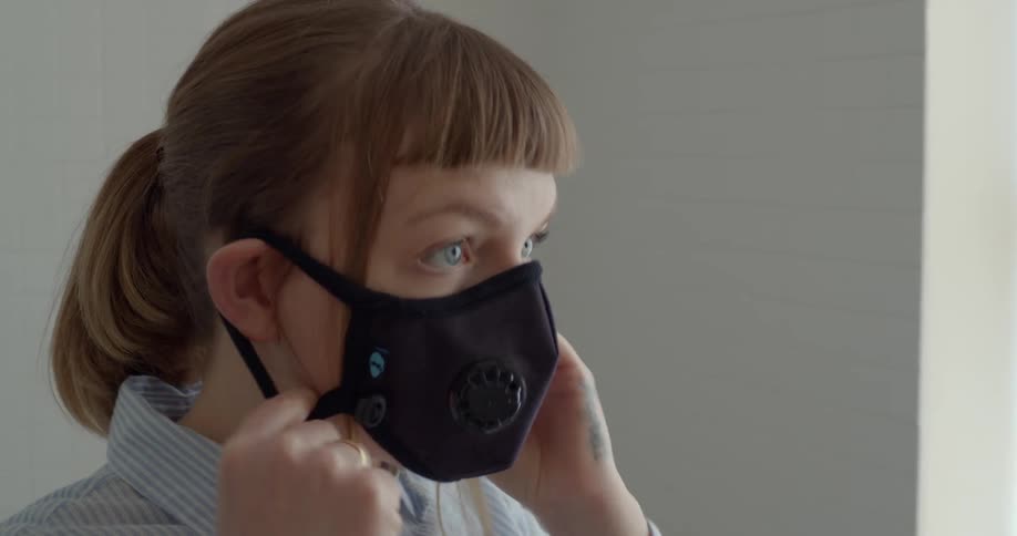 A white woman stands in a room and puts a face mask on