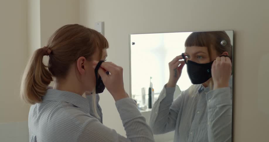 A white woman stands in front of a mirror and puts on a face mask