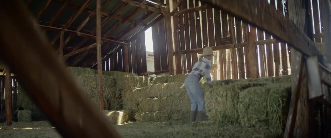 Blonde woman wearing a cowboy hat carries a bale of hay in the barn