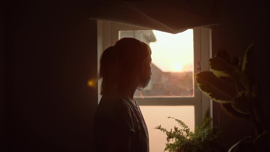 Silhouette of a woman wearing a cloth face mask looking out a window