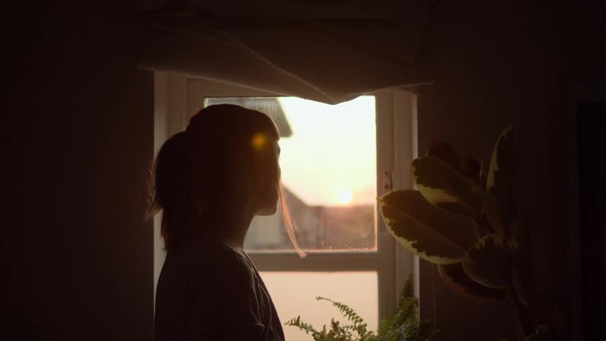 Woman looks out window toward sun rising on the horizon