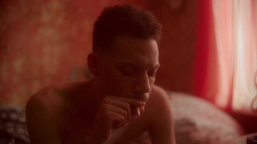 Man sitting on the bed breathes smoke into his mouth from a cigarette in his room