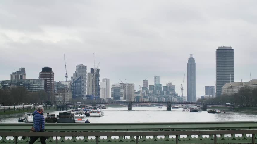 Two people walk across a pedestrian bridge by a large city and river