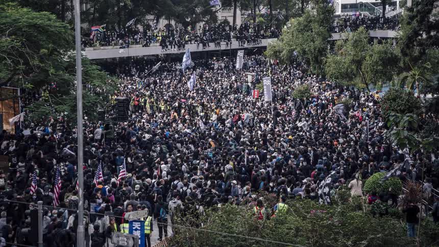 A large crowd of protesters gather and walk in a city park carrying banners and flags